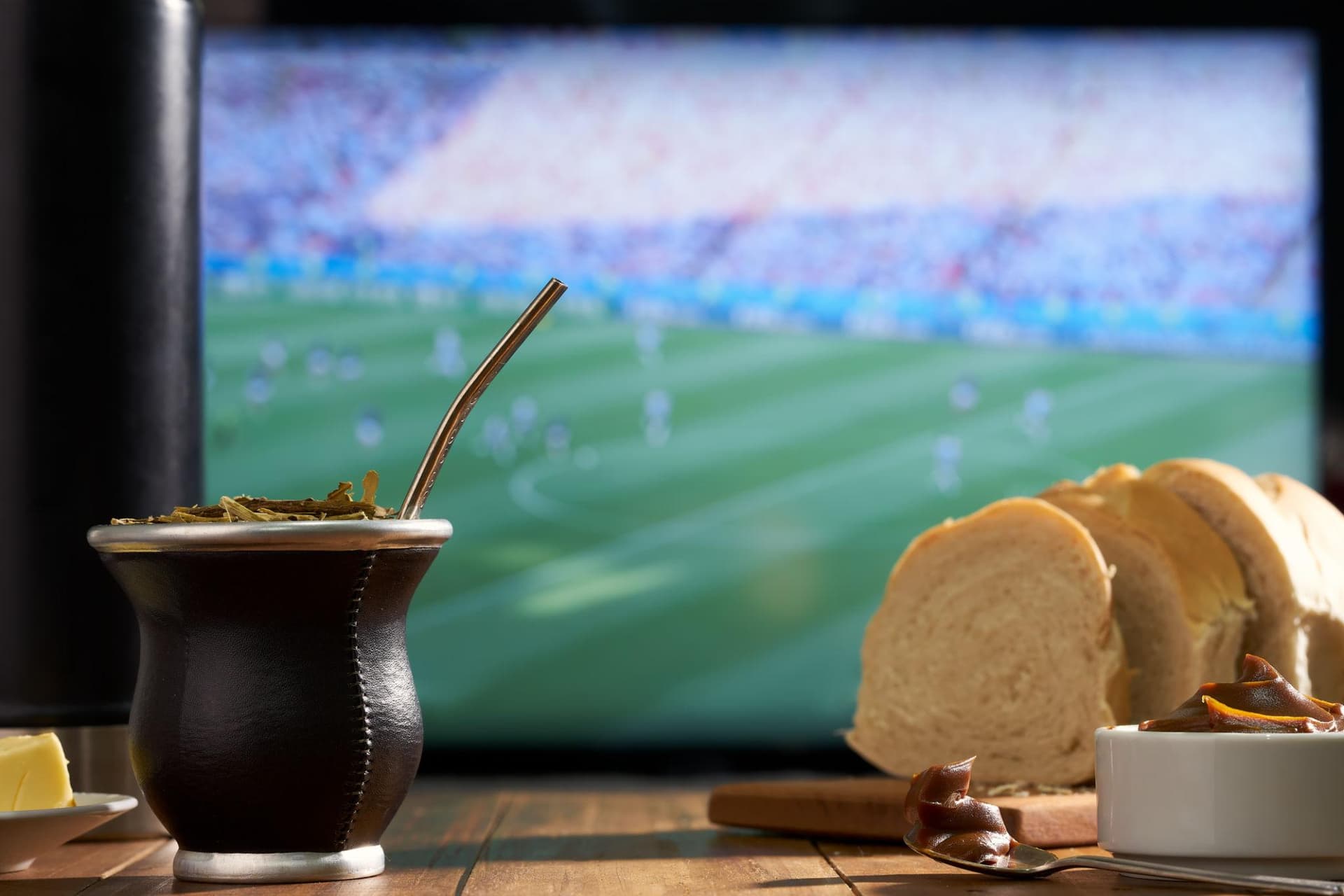 Mate argentino con bombilla frente a TV con partido de fútbol, pan y dulce de leche