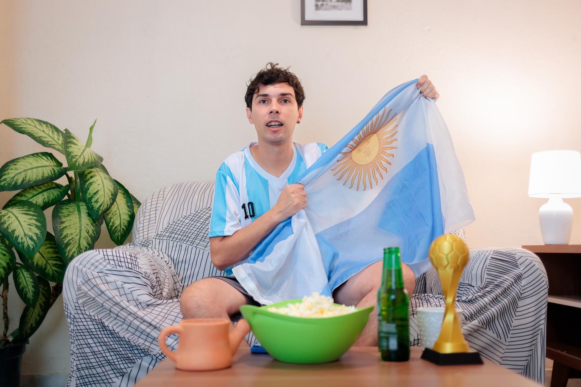 Hincha argentino con camiseta y bandera viendo partido en casa con pochoclo y cerveza